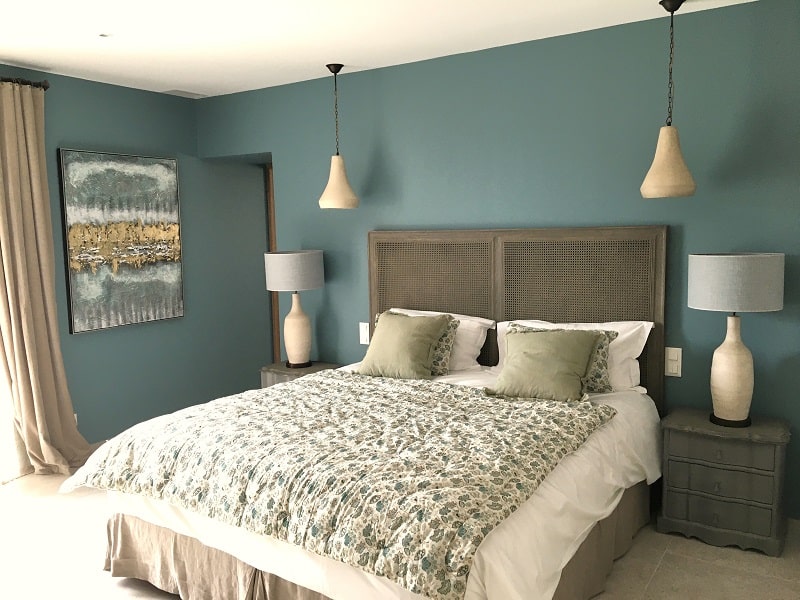Chambre moderne aux murs bleu canard. Grand lit, tête de lit rotin, boutis fleuri, lampes et tableau abstrait or.