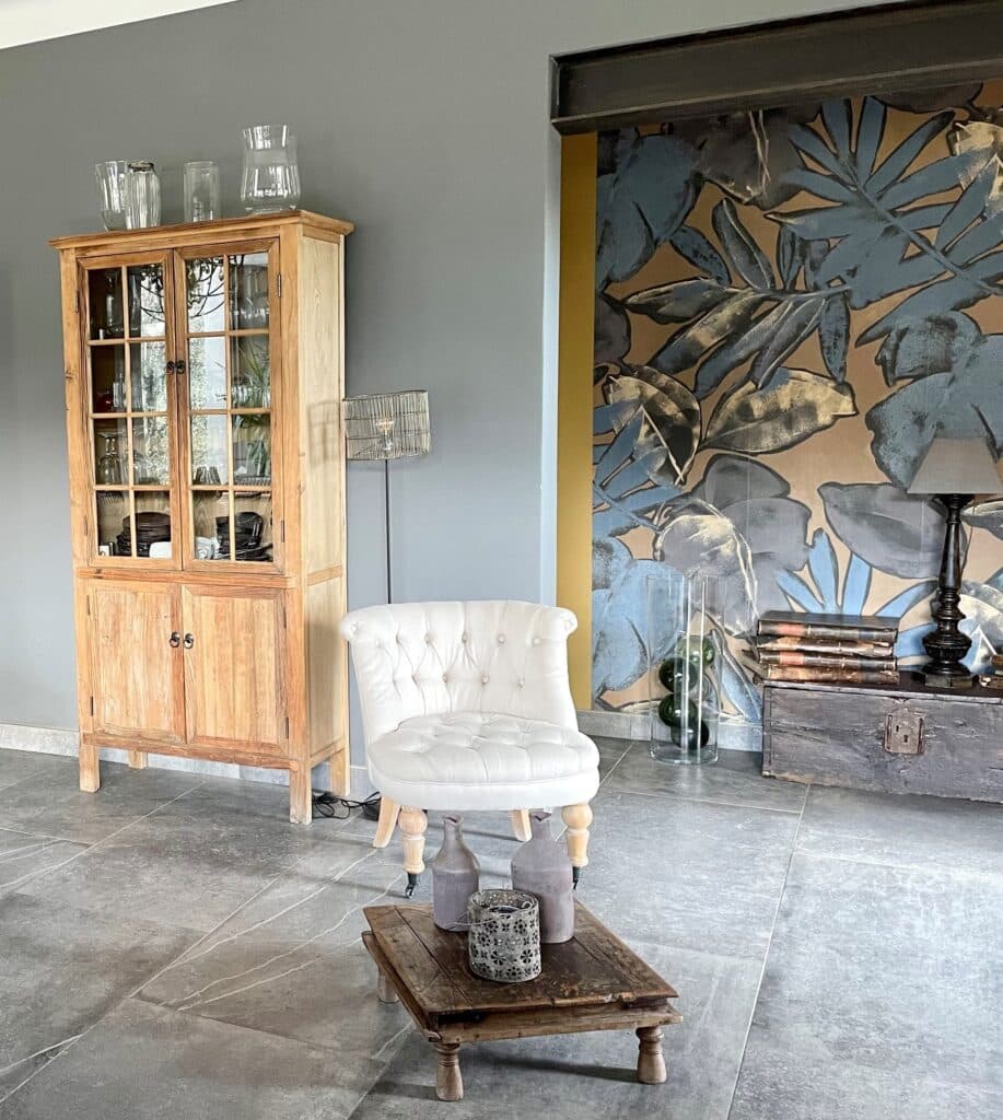 Intérieur chaleureux avec vitrine en bois clair, chaise capitonnée blanche, table basse rustique, et mur décoré de grandes feuilles bleues. Coffre ancien, lampe.