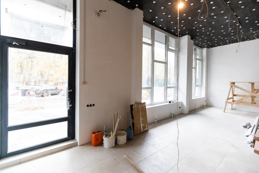 Intérieur en rénovation : pièce avec grandes fenêtres, murs clairs, plafond isolé noir, carrelage. Matériaux et fils apparents.